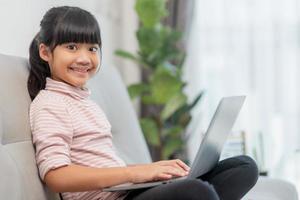 une petite écolière asiatique utilise un ordinateur portable assis sur un canapé seul à la maison. enfant apprenant à lire le contenu des médias sociaux en ligne, jouer à des cours d'éducation en discutant avec des amis. photo