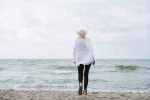 portrait d'une élégante femme blonde en chemise blanche sur une plage de sable à la mer de tempête au vent photo