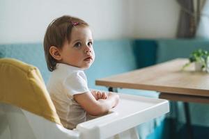 charmante petite fille assise dans une chaise haute pour se nourrir dans la cuisine lumineuse à la maison photo