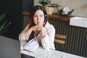 femme brune adulte de cinquante ans dans un casque faisant des notes dans un livre quotidien avec un ordinateur portable ouvert, étudie quelque chose avec un cours en ligne à la maison photo