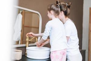 jeune mère femme aux cheveux longs avec une petite fille fille en pyjama se lavant le visage et les mains le matin à la maison, la famille passe du temps ensemble dans la salle de bain photo