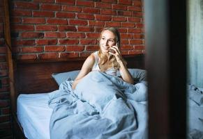 Femme adulte de quarante ans avec de longs cheveux blonds en pyjama à l'aide de téléphone portable assis sur le lit dans la chambre loft à la maison photo