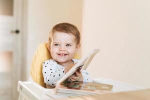 Happy cute baby girl in booster seat lecture livre pour enfants à la maison photo