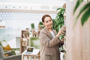 adulte femme d'affaires brune souriante de quarante ans aux cheveux longs en costume beige élégant et jeans dans un lieu public, bureau d'espace ouvert vert, coworking. enseignant ou mentor sympathique avec un livre à la bibliothèque photo