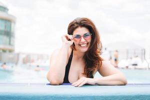 élégante jeune femme heureuse plus corps de taille positive en maillot de bain noir et lunettes de soleil profitant de sa vie dans la piscine de l'hôtel, vacances d'été photo