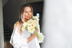 jeune belle femme souriante de quarante ans aux cheveux longs blonds en chemise blanche avec bouquet de fleurs jaunes dans les mains près de la fenêtre dans un intérieur lumineux à la maison photo