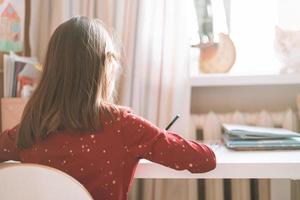 jolie écolière aux cheveux longs en robe rouge dessine avec un crayon à table dans la chambre des enfants à la maison photo