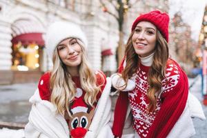 jeunes amies heureuses aux cheveux bouclés en rouge s'amusant dans la rue d'hiver décorée de lumières