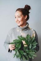 adulte belle femme souriante de quarante ans avec des cheveux bouclés brune en robe tricotée gris chaud avec une guirlande de noël sapin bricolage à la main sur le fond gris photo