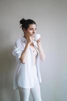 belle femme souriante adolescente étudiante aux cheveux longs noirs dans des vêtements de maison avec une tasse de café ou de thé le matin photo