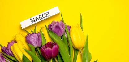 bannière de printemps. un bouquet de tulipes jaunes, roses et violettes et mars écrit sur un bloc de bois. concept de la journée de la femme photo