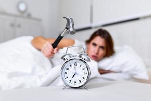 une jeune femme essaie de casser le réveil avec un marteau, de détruire l'horloge. fille allongée dans son lit éteignant un réveil avec un marteau le matin à 7h. photo