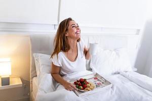 jeune femme souriante prenant un délicieux petit déjeuner et café sur un plateau au lit. femme prenant son petit déjeuner au lit. grand sourire à pleines dents. photo