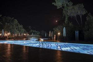 passerelle au-dessus de la piscine illuminée entourée de parasols et d'arbres la nuit photo