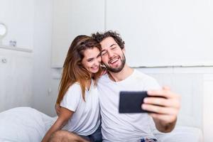 jeune couple au lit prenant selfie sur téléphone portable, jeune beau couple heureux sur le lit faisant autoportrait par smartphone et étreignant, photo