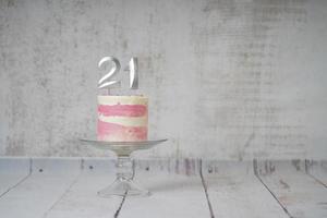 21ème gâteau d'anniversaire gâteau rose et argenté avec quelques pépites et 21ème chandelle sur un fond en bois blanc. photo