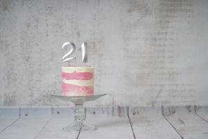 21ème gâteau d'anniversaire gâteau rose et argenté avec quelques pépites et 21ème chandelle sur un fond en bois blanc. photo