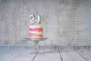 21ème gâteau d'anniversaire gâteau rose et argenté avec quelques pépites et 21ème chandelle sur un fond en bois blanc. photo