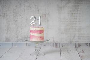 21ème gâteau d'anniversaire gâteau rose et argenté avec quelques pépites et 21ème chandelle sur un fond en bois blanc. photo