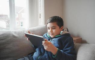 concept d'éducation, écolier tenant une tablette lisant un livre électronique pour les devoirs, portrait enfant heureux jouant au jeu en ligne sur internet avec des amis, jeune garçon regardant un dessin animé sur une tablette numérique, enfant assis sur un canapé photo