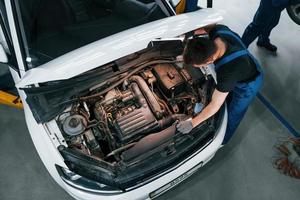 sous la capuche. un homme en uniforme de travail répare une automobile blanche à l'intérieur. conception de service automobile photo