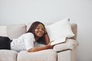 femme afro-américaine positive en chemise blanche allongée sur un canapé avec un bloc-notes dans les mains photo