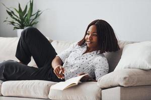 femme afro-américaine positive en chemise blanche allongée sur un canapé avec un bloc-notes dans les mains photo