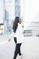 avec sac à provisions. jeune femme afro-américaine en chemise blanche à l'extérieur de la ville contre le bâtiment de l'entreprise photo