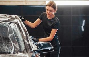 essuie le véhicule qui est au savon blanc. une automobile noire moderne est nettoyée par une femme à l'intérieur d'une station de lavage de voiture photo