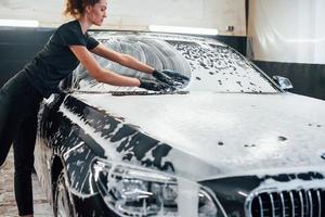 essuie le véhicule qui est au savon blanc. une automobile noire moderne est nettoyée par une femme à l'intérieur d'une station de lavage de voiture photo