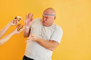 les mains avec des hamburgers tentent le gars. drôle d'homme en surpoids en cravate de tête sportive est sur fond jaune photo