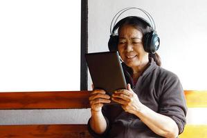 une femme senior asiatique heureuse porte des écouteurs, tient une tablette intelligente, écoute de la musique. concept, appareil intelligent de technologie d'utilisation des personnes âgées. détendez-vous avec de la musique en ligne, regardez des vidéos. apprendre de nouvelles choses sur Internet. photo