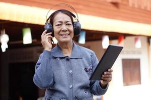 une femme senior asiatique heureuse porte des écouteurs, tient une tablette intelligente, écoute de la musique. concept, appareil intelligent de technologie d'utilisation des personnes âgées. détendez-vous avec de la musique en ligne, regardez des vidéos. apprendre de nouvelles choses sur Internet. photo