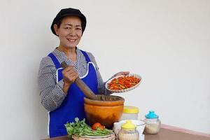 une femme senior asiatique heureuse cuisine, porte une casquette et un tablier de chef, tient un pilon, un mortier et une assiette de piments. concept, cuisiner pour la famille. style de vie de la cuisine thaïlandaise. activité des personnes âgées. photo