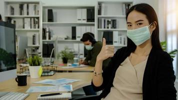 femme d'affaires portant un masque assis au bureau et montrant le pouce vers le haut