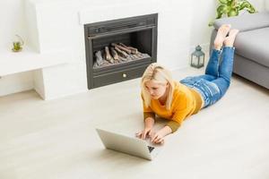 profiter de temps insouciants à la maison. jolie jeune femme travaillant sur un ordinateur portable et souriant en position couchée sur le tapis à la maison photo