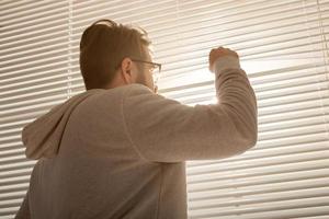 vue arrière du jeune homme élégant regardant à travers le trou dans les stores et regardant dans la rue. concept de profiter du soleil du matin et de la positivité photo