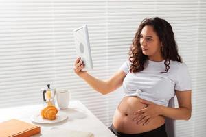 heureuse jeune femme enceinte belle parler à maman en utilisant l'appel vidéo pendant le petit déjeuner du matin. concept de communication et d'attitude positive pendant la grossesse photo