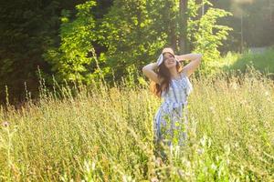 heureuse jeune femme aux cheveux longs en chapeau et robe marchant à travers la forêt d'été par une journée ensoleillée. concept de joie d'été photo