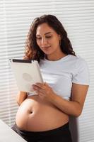 femme enceinte hispanique utilisant une tablette numérique pendant le petit-déjeuner. technologie, grossesse et congé de maternité photo