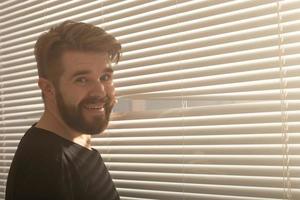 un jeune homme à la barbe regarde à travers le trou dans les stores et regarde dans la rue. concept de surveillance et de curiosité photo