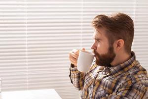 vue latérale d'un jeune homme d'affaires barbu pensif buvant une tasse de café sur fond de stores. concept de matinée agréable ou de pause déjeuner. fond photo