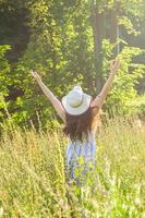 vue arrière d'une jeune femme heureuse aux cheveux longs en chapeau et robe tire ses bras tout en marchant à travers la forêt d'été par une journée ensoleillée. notion de joie de vivre photo
