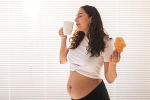belle femme enceinte tenant un croissant et une tasse de café dans ses mains pendant le petit déjeuner du matin. concept de bonne santé et d'attitude positive en attendant bébé. espace de copie.