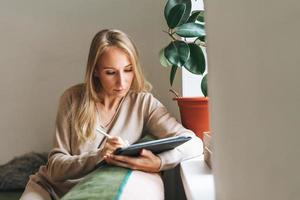 belle jeune femme blonde artiste illustratrice dessinant sur une tablette près de la fenêtre à la maison photo