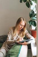 belle jeune femme blonde artiste illustratrice dessinant sur une tablette près de la fenêtre à la maison photo
