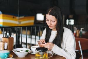 séduisante jeune femme brune souriante en mode décontracté utilisant un téléphone portable prenant un brunch au café photo