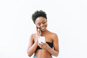 souriante belle jeune femme afro-américaine en haut noir à l'aide d'un téléphone portable isolé sur fond blanc photo