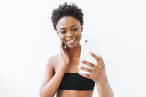 souriante belle jeune femme afro-américaine en haut noir à l'aide d'un téléphone portable isolé sur fond blanc photo