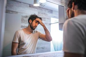 beau jeune homme touchant ses cheveux avec la main et se toilettant dans la salle de bain à la maison. homme métrosexuel blanc inquiet pour la perte de cheveux et regardant le miroir de sa chute de cheveux. photo